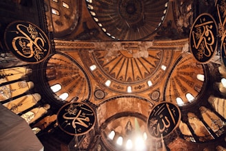An intricate, ornately decorated dome with detailed Islamic calligraphy and large circular arabesque medallions against a backdrop of golden and earthy tones. Light pours in through small windows, illuminating the intricate patterns on the structure.