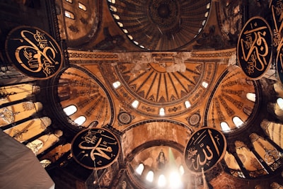 An intricate, ornately decorated dome with detailed Islamic calligraphy and large circular arabesque medallions against a backdrop of golden and earthy tones. Light pours in through small windows, illuminating the intricate patterns on the structure.