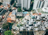 Aerial view of a cityscape featuring high-rise buildings with unique architecture, characterized by a staggered facade with balconies and rooftop gardens. Surrounding the buildings are streets bustling with traffic and smaller structures with red-tiled roofs. Lush greenery and trees are interspersed among the buildings, adding a touch of nature to the urban environment.
