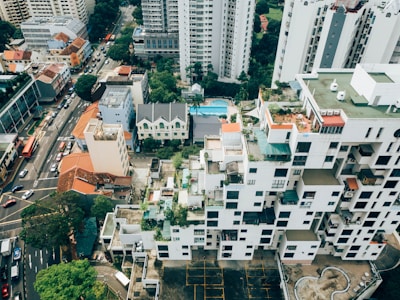 Aerial view of a cityscape featuring high-rise buildings with unique architecture, characterized by a staggered facade with balconies and rooftop gardens. Surrounding the buildings are streets bustling with traffic and smaller structures with red-tiled roofs. Lush greenery and trees are interspersed among the buildings, adding a touch of nature to the urban environment.