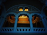 An architectural interior featuring an ornate balcony with curved archways and decorative columns. A chandelier is hanging from the ceiling in the background, casting a warm light that contrasts with the cool blue tone of the structure. The design elements suggest a classical style.