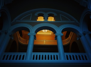 An architectural interior featuring an ornate balcony with curved archways and decorative columns. A chandelier is hanging from the ceiling in the background, casting a warm light that contrasts with the cool blue tone of the structure. The design elements suggest a classical style.