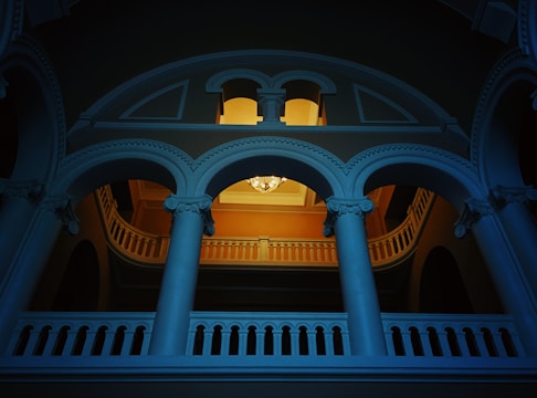 An architectural interior featuring an ornate balcony with curved archways and decorative columns. A chandelier is hanging from the ceiling in the background, casting a warm light that contrasts with the cool blue tone of the structure. The design elements suggest a classical style.