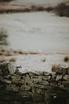 A rustic stone wall made of irregularly shaped rocks stands prominently in the foreground. The stones are stacked with varying sizes and shades of gray and beige. The background is an expansive, blurred landscape, suggesting a rural or pastoral setting with muted, earthy tones.