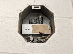An octagonal electrical box installed into a wall, containing various electrical components including a light bulb, coils, and a circuit diagram. The box is metallic and surrounded by textured white plaster or concrete.