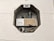 An octagonal electrical box installed into a wall, containing various electrical components including a light bulb, coils, and a circuit diagram. The box is metallic and surrounded by textured white plaster or concrete.