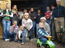 A group of people of various ages are gathered together, appearing to be at a casual outdoor event. Some are holding drinks, and a young child in front is sitting on a toy car. The setting looks like a backyard with wooden structures, possibly a shed or garage.