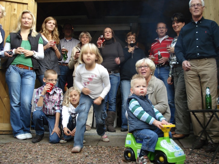 A group of people of various ages are gathered together, appearing to be at a casual outdoor event. Some are holding drinks, and a young child in front is sitting on a toy car. The setting looks like a backyard with wooden structures, possibly a shed or garage.