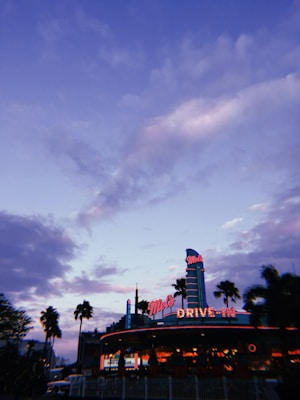 A retro diner with neon lights is set against a dramatic evening sky filled with clouds. Palm trees frame the scene, adding a tropical feel. The architecture and signage exude a mid-20th-century American aesthetic.