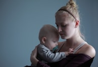 A woman is holding a baby closely, creating an intimate, tender moment. The baby is resting its head on her shoulder, and she looks contemplative with her eyes closed. The background is a solid muted blue, enhancing the calm atmosphere.