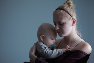 A woman is holding a baby closely, creating an intimate, tender moment. The baby is resting its head on her shoulder, and she looks contemplative with her eyes closed. The background is a solid muted blue, enhancing the calm atmosphere.