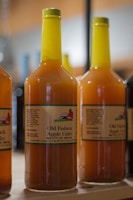 Several bottles of apple cider with yellow caps are arranged on a wooden shelf. The labels feature a drawing of a barn and contain text reading 'Old Fashion Apple Cider.'