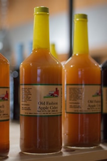 Several bottles of apple cider with yellow caps are arranged on a wooden shelf. The labels feature a drawing of a barn and contain text reading 'Old Fashion Apple Cider.'