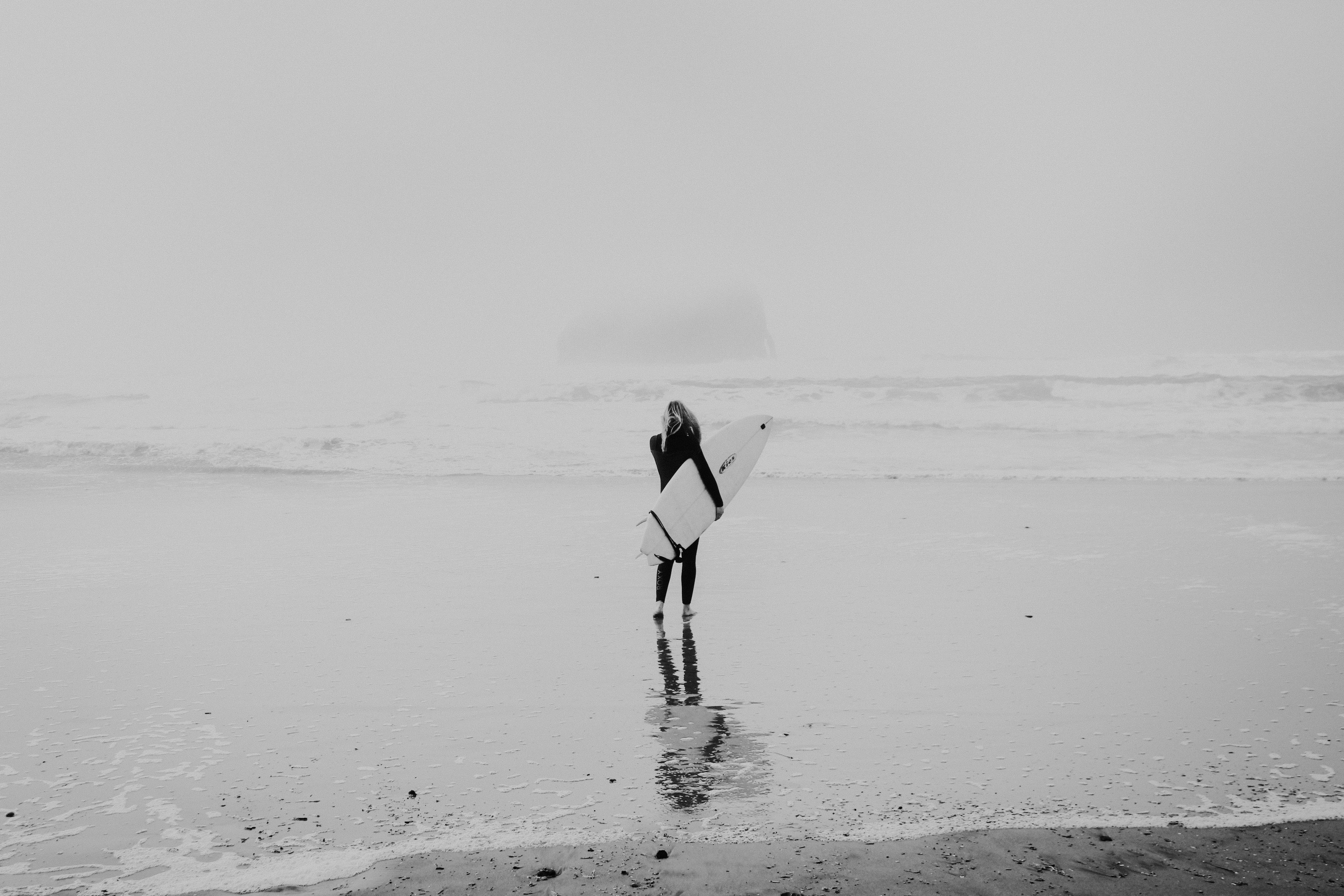 grayscale photo of woman walking on seashore while carrying surfboard