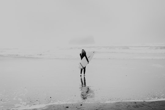 grayscale photo of woman walking on seashore while carrying surfboard in Cape Kiwanda United States