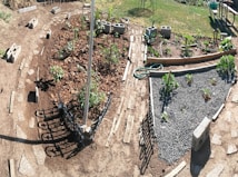 A garden with various sections of planted vegetables and plants, bordered by wooden planks and concrete blocks. There are pathways of stone leading between the plots, and a hose is coiled on the ground. A picnic table and chairs are visible in the background on a grassy area.