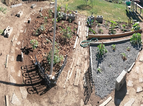 A garden with various sections of planted vegetables and plants, bordered by wooden planks and concrete blocks. There are pathways of stone leading between the plots, and a hose is coiled on the ground. A picnic table and chairs are visible in the background on a grassy area.