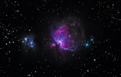 A vibrant space scene featuring a nebula with bright pink and purple hues in the center against a backdrop of numerous stars scattered across a black sky. A bright blue star is visible on the right side, while another smaller cluster of nebulae is seen on the left.