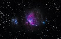A vibrant space scene featuring a nebula with bright pink and purple hues in the center against a backdrop of numerous stars scattered across a black sky. A bright blue star is visible on the right side, while another smaller cluster of nebulae is seen on the left.