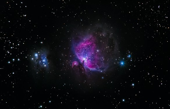 A vibrant space scene featuring a nebula with bright pink and purple hues in the center against a backdrop of numerous stars scattered across a black sky. A bright blue star is visible on the right side, while another smaller cluster of nebulae is seen on the left.