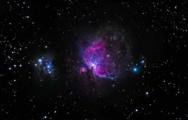 A vibrant space scene featuring a nebula with bright pink and purple hues in the center against a backdrop of numerous stars scattered across a black sky. A bright blue star is visible on the right side, while another smaller cluster of nebulae is seen on the left.