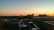 A rooftop view of a cityscape with various buildings silhouetted against the backdrop of a sunset. The sky transitions from blue to orange, casting a warm glow over the scene. Rooftop HVAC units and pipes are visible in the foreground.