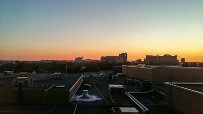 A rooftop view of a cityscape with various buildings silhouetted against the backdrop of a sunset. The sky transitions from blue to orange, casting a warm glow over the scene. Rooftop HVAC units and pipes are visible in the foreground.