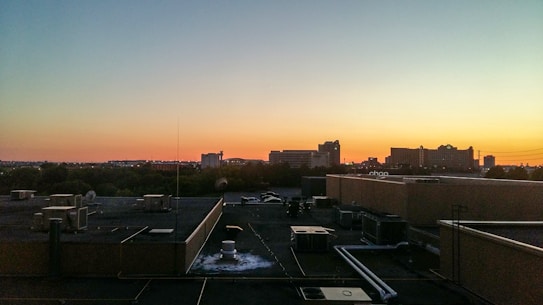 A rooftop view of a cityscape with various buildings silhouetted against the backdrop of a sunset. The sky transitions from blue to orange, casting a warm glow over the scene. Rooftop HVAC units and pipes are visible in the foreground.