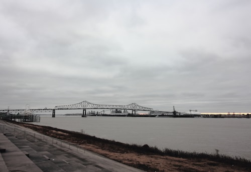 A large steel bridge spans across a wide river. The bridge has a truss design and is supported by several pillars. In the background, there is an industrial area with cranes and buildings. The sky is overcast with thick, grey clouds. The foreground features a broad riverside walkway with paved tiles and a railing, adjacent to a strip of vegetation.