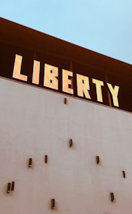 A large building facade with the word 'LIBERTY' prominently displayed in bold, glowing letters at the top. Below the sign, the wall is plain with several small, protruding fixtures evenly spaced out.