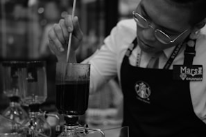 A person wearing glasses and an apron is seen stirring a dark liquid in a glass apparatus. Several other similar glass apparatuses can be seen in the background, indicating a scientific or culinary setting, possibly related to coffee brewing.