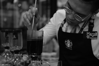 A person wearing glasses and an apron is seen stirring a dark liquid in a glass apparatus. Several other similar glass apparatuses can be seen in the background, indicating a scientific or culinary setting, possibly related to coffee brewing.
