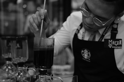 A person wearing glasses and an apron is seen stirring a dark liquid in a glass apparatus. Several other similar glass apparatuses can be seen in the background, indicating a scientific or culinary setting, possibly related to coffee brewing.