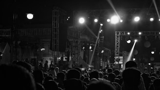 A monochrome image depicting a large crowd gathered at a nighttime event. Bright spotlights illuminate a stage with scaffolding and lighting equipment, creating stark contrasts and dramatic shadows. Silhouettes of people are visible in the foreground, watching the performance with anticipation.