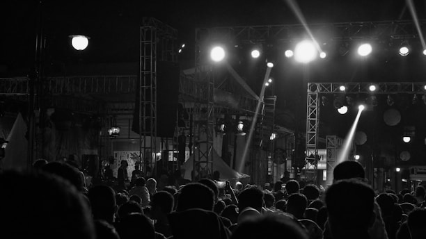 A monochrome image depicting a large crowd gathered at a nighttime event. Bright spotlights illuminate a stage with scaffolding and lighting equipment, creating stark contrasts and dramatic shadows. Silhouettes of people are visible in the foreground, watching the performance with anticipation.