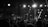 A monochrome image depicting a large crowd gathered at a nighttime event. Bright spotlights illuminate a stage with scaffolding and lighting equipment, creating stark contrasts and dramatic shadows. Silhouettes of people are visible in the foreground, watching the performance with anticipation.