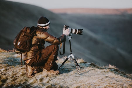 A person wearing a winter jacket and beanie is crouched down on a grassy hill, focusing on taking a photograph with a camera mounted on a tripod. The setting appears to be a scenic outdoor landscape, possibly at sunrise or sunset.