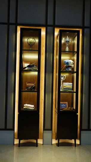 A pair of illuminated wooden display cabinets contain various items, including a decorative globe, a trophy, folded clothes, shoes, and framed pictures. The arrangement is neat and organized with some items showcasing luxury and elegance.