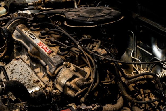 An engine compartment showcasing various components, including a Toyota engine with 16 valves. The image highlights the complex arrangement of hoses, wires, and metal parts, exhibiting signs of use and exposure to dirt and grime.