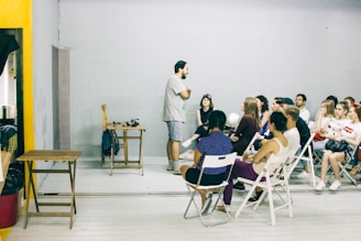 A group of people is seated in a room with minimalistic decor. One person stands at the front, engaging in conversation or giving a presentation. The ambiance is informal, and the participants appear attentive and relaxed. Foldable chairs are used for seating, and a small table is placed at the front with some items on it.