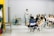 A group of people is seated in a room with minimalistic decor. One person stands at the front, engaging in conversation or giving a presentation. The ambiance is informal, and the participants appear attentive and relaxed. Foldable chairs are used for seating, and a small table is placed at the front with some items on it.