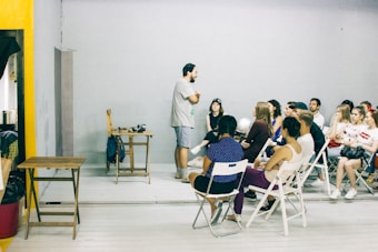 A group of people is seated in a room with minimalistic decor. One person stands at the front, engaging in conversation or giving a presentation. The ambiance is informal, and the participants appear attentive and relaxed. Foldable chairs are used for seating, and a small table is placed at the front with some items on it.