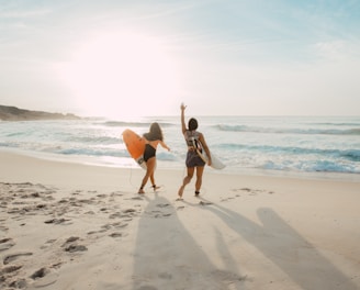 two women walking towards the ocean carrying surfboards during day
