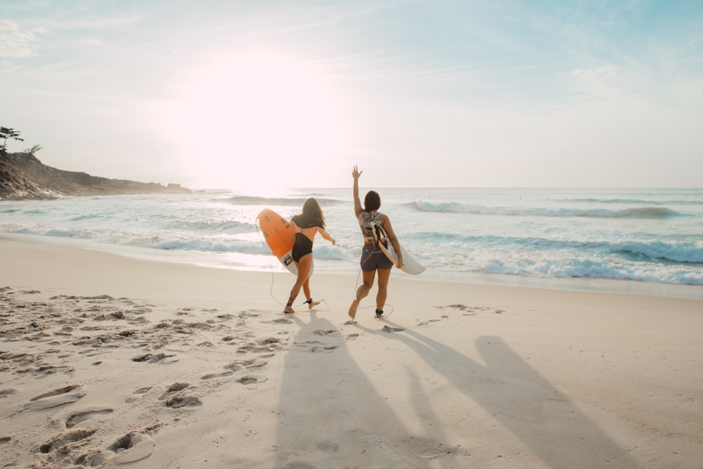 two women walking towards the ocean carrying surfboards during day