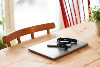 A minimalist wooden table holds a closed laptop, a smartphone, and a smartwatch placed on top. The background includes a red chair and a small potted plant, adding a touch of greenery to the setting.