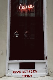 A window display features a neon sign with the word 'love' in pink, casting a glow on three reflective skull objects positioned against the dark backdrop. Below the window, the phrase 'LOVE LETTERS ONLY' is painted in red on a white surface.