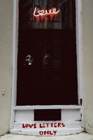 A window display features a neon sign with the word 'love' in pink, casting a glow on three reflective skull objects positioned against the dark backdrop. Below the window, the phrase 'LOVE LETTERS ONLY' is painted in red on a white surface.