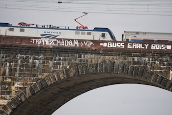 A train labeled with the Amtrak logo travels over a stone bridge. The bridge is marked with graffiti containing white spray-painted words, and there are overhead electrical lines visible.