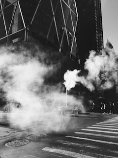 A black and white urban scene featuring a large amount of steam rising from a vent in the street. The background is dominated by a tall, modern building with crisscrossing structural elements. The foreground includes a crosswalk and several pedestrians, partially obscured by the steam, walking along the sidewalk.