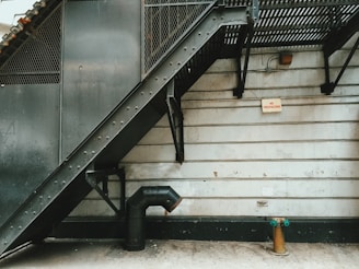 A metal fire escape staircase descends along the side of a concrete building. The structure features industrial design elements, such as rivets and metal mesh. A pipe extends from the wall near the ground, next to a fire hydrant with green caps. A 'No Trespassing' sign is mounted on the wall.